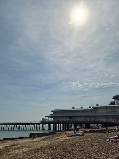The image depicts a sunny day at Felixstowe, United Kingdom. The sky is light blue with wispy clouds and the sun shining brightly. The "Boardwalk Cafe" pier structure is visible on the right. People are relaxing on the pebble beach along the edge of the pier. A man in blue swim trunks is lying on the beach, while other individuals are standing or sitting nearby. Birds are flying in the sky. The pier extends over the water. A woman is sitting on the sand further along the beach.