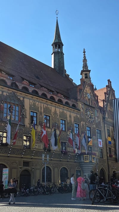 The image captures a sunlit view of the Ulm City Hall (Rathaus Ulm) in Ulm, Germany. The ornate facade of the building is adorned with vibrant murals and decorated with numerous flags, including those of Denmark, Italy, and Ukraine, among others. A large clock face is prominently displayed on the building's facade. To the left, a slender, green-patinated spire with a decorative weather vane rises against a clear blue sky. To the right, another, more intricate spire is visible. In the foreground, a cobblestone plaza is populated by several people and a rack of parked bicycles. A woman in the foreground is walking with a black case. Other individuals are standing and conversing, one woman wearing a bright pink dress. The overall scene depicts a lively urban setting on a clear day.