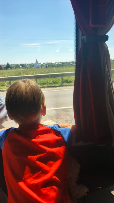 A young boy with light brown hair, seen from behind, sits by a bus window, looking out at a landscape that appears to be the outskirts of Sokilnyky, Ukraine. The boy wears a red t-shirt with blue sleeves. He is holding a fuzzy white toy. Through the window, a rural scene is visible, including a road, green fields, and a town with a white building, possibly a church, with a dome. A red curtain partially obscures the right side of the window. Sunlight casts shadows across the boy's back.