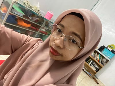 The image is a close-up, angled selfie of a woman wearing a pink hijab and eyeglasses. She has dark hair visible under the hijab, dark eyes, and is wearing red lipstick. The background shows two shelves filled with various items. The shelf on the left appears to contain folded fabrics in bright colors like orange and green, along with other unidentifiable objects. The shelf on the right is filled with books, folders, and other miscellaneous items, including a blue box with "natural" written on it and a yellow bag. The woman is wearing a pink top that drapes over her arm. The environment appears to be an indoor setting, possibly a room with tiled flooring.