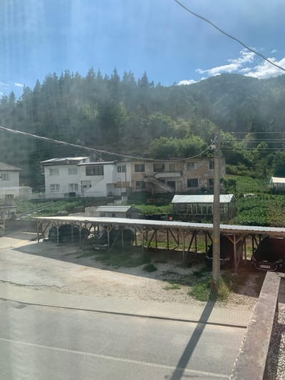The image depicts a village scene in Krushev Dol, Bulgaria, with a mountainous backdrop covered in dense green forest. A clear blue sky with scattered white clouds suggests it is daytime with fair weather. The foreground features a road with a paved sidewalk and a gravel area. To the left, there's a series of modern white houses nestled against the hillside, interspersed with some older, weathered buildings, some of which appear to be under renovation or in disrepair. Several greenhouses are visible among the houses and vegetation.
On the right side of the image, a covered parking area with a corrugated metal roof is supported by a rustic wooden structure. Several dark-colored cars are parked underneath it, including one with a license plate visible. A utility pole with wires stretches across the scene, a common sight in rural areas. The overall impression is of a lived-in, working village, perhaps with some tourism or agricultural activities given the greenhouses. The image composition, with the road and covered parking in the foreground and the houses and natural landscape behind, creates a sense of depth and perspective. There is no discernible emotional tone, and the focus is on documenting the environment.