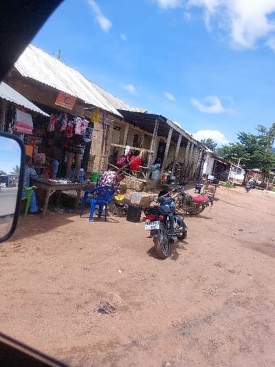 The image depicts a commercial street in Kimamba, Tanzania, under a clear blue sky with scattered white cumulus clouds. The foreground and mid-ground consist of an unpaved, reddish-brown dirt surface, exhibiting tire tracks and minor debris.
On the right side of the scene, a series of connected single-story structures with corrugated metal roofs serve as shops or market stalls. Their fronts are open, displaying various goods. Clothing items, including dresses and shirts of assorted colors and patterns, hang on racks and are draped over tables. Other merchandise, such as plastic containers and bags, is also visible.
Multiple individuals are present. On the far left, a person in a patterned shirt stands near a wooden table. A green plastic chair is partially visible next to this table. Further into the shops, another individual sits on a raised platform or bench. Other figures are indistinctly visible inside or near the shop entrances.
Two motorcycles are parked on the dirt surface. A dark-colored motorcycle, bearing the license plate T 890 BZ, is positioned in the mid-ground, facing away from the viewer. Behind it, a second motorcycle, colored red and green, is also parked. A blue plastic chair and a yellow plastic basin are situated near the shop fronts. The lower-left corner of the image contains the reflection of a vehicle's side mirror. Trees are visible in the distant background on the right side of the frame.