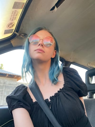 A young woman with blue hair is sitting in the passenger seat of a car, looking up and to the right. She is wearing round, gold-rimmed sunglasses with pink lenses that reflect the sky and buildings. Her black top has puffed sleeves and a gathered neckline. A black seatbelt is visible across her chest. The car's interior is visible, including the gray fabric of the car seat and the headliner. On the headliner, there are stickers, including one with a circular graphic and text that appears to be in a Cyrillic script. Outside the car window, a building with a tiled roof and a concrete wall can be seen. The sun is shining brightly, suggesting it is daytime. The overall scene depicts a candid moment of a woman in a vehicle, likely on a journey.