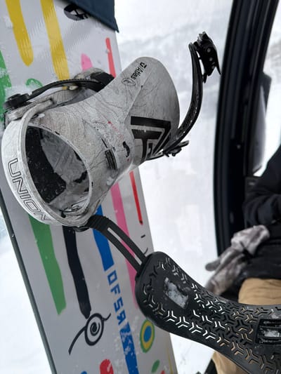 A close-up shows a snowboard with Union Atlas bindings. The bindings are silver with a textured pattern. One binding is partially open. The snowboard has colorful horizontal stripes of pink, blue, green, and orange, along with a black spiral design. The letters "DFSR" are visible between the stripes. The background shows snow and a dark, blurry figure.
