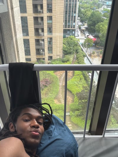 A person with dreadlocks is lying down next to a window with a view of Binjiang District, China. The view shows a building with multiple windows and air conditioning units attached, plus a garden area with pathways. The weather appears to be overcast.