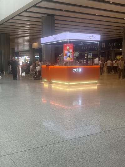 In this vertical, outdoor photograph taken in Parakar, Armenia, a bright orange kiosk with the "cofix" logo is prominently displayed. The kiosk features an illuminated sign with "cofix" written in white, and another sign on the side also says "cofix new mojitos" with images of drinks. People are seen standing and sitting around the kiosk, suggesting it's an active place, possibly a cafe or refreshment stand. In the background, there's a sign with "Arriva" and "ժամանում," indicating an arrivals area, likely an airport or transportation hub. The floor is tiled and reflects the lights from the kiosk, adding a dynamic element to the scene. The overall atmosphere appears to be busy and functional, with the lighting suggesting it could be daytime or early evening. The visible text includes brand names and Armenian script, adding a touch of local context to the international setting.