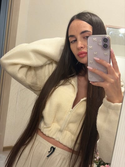 The image is a mirror selfie showing a young woman indoors. She has long, straight, dark brown hair that flows over her shoulders and chest. She is wearing a cream or off-white outfit, consisting of a cropped, long-sleeved jacket made of a soft, fuzzy, fleece-like material, featuring a white zipper down the front. Matching ribbed pants or shorts are partially visible on her lower right side, with a small dark emblem or design near the waist.
Her left hand is holding a smartphone, positioned in the upper central part of the image, capturing her reflection. Her fingernails are long, almond-shaped, and painted a light pink. The smartphone case is a distinct lavender or light purple color and is decorated with multiple small, white, flower-shaped embellishments.
The woman's face is visible, turned slightly towards the camera with her dark eyes looking out from under well-defined eyebrows. Her lips are full and slightly parted in a soft pout.
The background reflected in the mirror appears to be a plain, light-colored wall. In the upper right corner of the reflected space, there is a soft, out-of-focus hint of pink, which could be artificial flowers or a decorative element. The entire image is oriented diagonally, indicating it was taken at an angle or has been rotated. The lighting in the scene is soft and even, suggesting an indoor environment without harsh shadows.