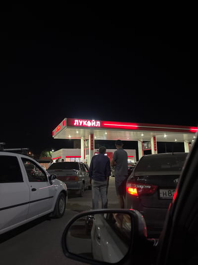 This media file captures a scene at a Lukoil gas station at night, with the image rotated 90 degrees counter-clockwise. Two men are visible, one wearing a grey t-shirt and a black cap, and the other in a dark shirt. They appear to be standing near a dark-colored car. Behind them, a white car is parked.
The gas station structure is illuminated with bright red neon lights, and the Lukoil logo is prominently displayed in large, glowing red letters. The sign also has Cyrillic text that reads "ЛУКОЙЛ". A car license plate is partially visible on the dark car, with text that appears to be "H 87". Gas pumps are visible in the background. The scene suggests people are at the gas station, possibly refueling or interacting. The lighting indicates it is nighttime, and the weather appears to be clear. The location is identified as Кирпичного завода № 1, Russia.