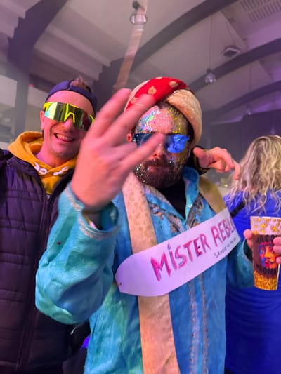 Two men and a woman are visible in the image. One man wears a yellow hoodie, a black puffer jacket, and neon yellow sunglasses. He smiles and holds up three fingers. The other man, slightly behind, sports a colorful turban and glitter on his face. He wears a blue and gold garment with a "Mister Rebul" sash. Part of a woman's blond hair and a box featuring a portrait are visible at the top. The overall setting appears to be a festive or celebratory event.