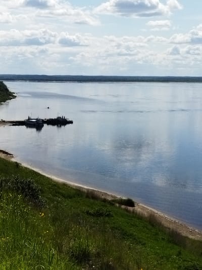 The image depicts a wide, calm river with a shoreline in the foreground. A grassy slope covered in green grass and some yellow wildflowers descends to a sandy beach, which then meets the water. A dock with two boats moored to it extends from the left side of the frame into the river. The water reflects the sky, which is partly cloudy with a mix of white and grey clouds. In the distance, a dense line of trees forms the opposite bank of the river, creating a horizontal division between the water and the sky. The overall scene is one of natural tranquility with a touch of human presence due to the dock and boats. The location is identified as Ershovka, Russia.