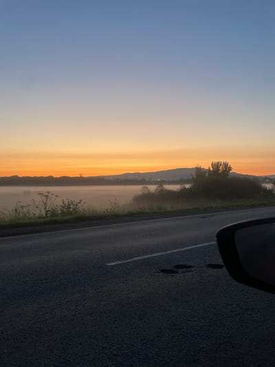 The image depicts a roadside view during a sunrise or sunset. The sky transitions from a deep blue at the top to a vibrant orange and yellow hue near the horizon. In the middle ground, a misty landscape is visible with silhouettes of trees and hills. A road, appearing to be asphalt, occupies the foreground, with a white line marking its edge. The side mirror of a vehicle is partially visible in the lower right corner. The scene conveys a serene, natural setting, possibly on the outskirts of a settlement. There is no specific identifiable information to suggest the location is the city of Uba, Russia.