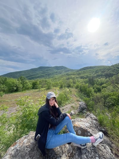 A young woman sits on a rocky outcrop, likely a cliff edge, with a valley and wooded hillside visible in the background. The sky is partly cloudy with bright sunlight. She wears light blue jeans, sneakers with pink socks, a black jacket, and a gray cap. She has dark hair and is looking toward the camera. The scene appears to be outdoors, potentially in a mountainous or hilly region.
While I do not know the exact location, based on the prompt I can infer that this scene is in the vicinity of Kamchia, Bulgaria, but I do not have enough information to pinpoint the specific spot.