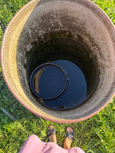 A first-person perspective shot looks down into an open, large, cylindrical concrete well filled with dark water. A black plastic bucket floats in the water. The inner rim of the well is weathered and discolored. The exterior of the well is partially visible, showing a reddish-brown rim. Green grass surrounds the well, and two bare feet wearing black sandals are visible in the foreground. The scene appears to be outdoors in daylight, possibly in Verkhneyarkeyevo, Russia, given the context provided.