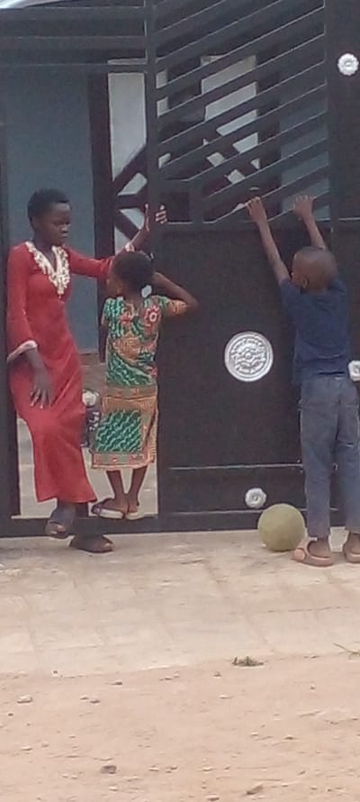 Three individuals are present in an outdoor scene in Agunmo, Nigeria. The background features a black metal gate with horizontal bars. An adult in a long red dress is lying on their back. Above them, a child in a green patterned dress is positioned with their head resting on their hand. A second child in a blue shirt and grey pants is also lying down, with their arms stretched out towards the gate. A green ball is located near the feet of the second child. The ground appears to be a dirt surface.