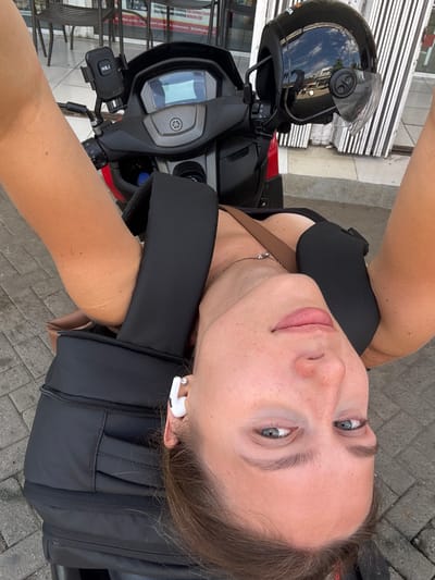 An upside-down, close-up, selfie-style, outdoor photograph depicts a woman on a scooter in Taliwang, Indonesia. The woman, who is the primary subject, is positioned in the lower half of the frame. Her head is tilted back and upside down, with her face looking towards the viewer. She has fair skin with freckles and light blue eyes. She is wearing white wireless earbuds, and a simple silver necklace. Her lips are slightly parted. Her arms are visible on either side of her head, bent at the elbows. She appears to be wearing a black top, with a brown strap visible across her chest.
The scooter is the secondary subject, positioned in the upper half of the frame. A black helmet with a clear visor is attached to the scooter's handlebars, reflecting the sky and surrounding environment. A black phone mount with the letters "HSJ" is also attached to the handlebars. The scooter's dashboard and front panel are visible.
The environment suggests an urban setting with a paved ground. In the background, there are tables and chairs, likely from an outdoor cafe or restaurant, with vertical striped awnings or shutters. The overall scene is brightly lit, indicating daylight. The image appears to be taken on a street or sidewalk area.