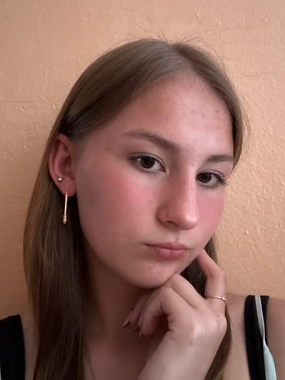 The media file contains a close-up, vertically oriented selfie of a young woman. She has light brown hair, fair skin with a rosy blush on her cheeks, and dark brown eyes. Her left hand is positioned near her face, touching her chin, and she is wearing a silver ring on her right index finger. She has a long, bar-shaped earring in her left ear, adorned with small stones, and a smaller stud earring in her right ear. A thin silver necklace is visible around her neck. She is wearing a striped top with black and light blue bands. The background appears to be a peach-colored wall with a textured surface. The lighting suggests an indoor setting, possibly during the daytime. Her expression is calm and contemplative. There is no visible text in the image. The location context provided is Qarabalıq, Kazakhstan.