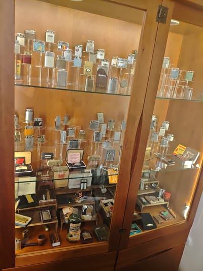 Within a wooden display cabinet featuring glass shelves and doors, numerous vintage lighters are meticulously arranged for exhibition in Swindon, United Kingdom. The cabinet's interior features a warm, light brown background and is illuminated by an internal light source on the right side.
The lighters, primarily constructed from various metals such as polished silver-tone, brass, and some with textured or enameled decorative panels (including a ship motif and abstract patterns), are displayed upright on clear acrylic risers and directly on glass shelves. Brands like "Ronson" are discernible on several lighters and their packaging.
Additional items on display include various Ronson brand boxes for flints and lighters, a box for "Zorro" lighters, a small green bottle labeled "The Celerity Lighter Fluid," a traditional wooden smoking pipe, and several other miscellaneous lighters, accessories, and promotional materials in various states of packaging. Small wooden blocks and a black case or pouch are also present on the lower shelves. No individuals are visible in the scene.