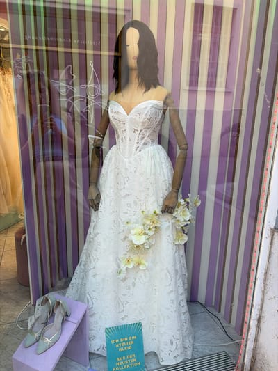A mannequin is displayed in a store window, wearing a white lace wedding dress. The mannequin has dark hair and is holding a bouquet of white flowers. In front of the mannequin, on a purple stand, is a pair of light blue high-heeled shoes with gold accents. A sign in the lower right corner reads "ICH BIN EIN ATELIER KLEID AUS DER NEUESTEN KOLLEKTION".

The background of the display features vertical stripes of purple and off-white. Reflections in the window include text that appears to read "Stadtbesichtigungen in Kimmelsbach" and a person holding a phone.

This storefront is located on Theaterstraße in Würzburg, Germany, indicated by the text visible in the window reflections.
