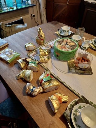The image shows a wooden table with various items arranged on it. There are cups and saucers with a green ivy pattern, a glass mug, a green tin with an image of people on it, and a clear container filled with marzipan balls. Several chocolate figures wrapped in gold foil, including bunnies and a horse head, are scattered across the table. There is a table runner with a decorative border. A glimpse of what appears to be a glass cabinet filled with items and a chair can be seen in the background. The presence of German food items suggests this is in Ganderkesee, Germany.