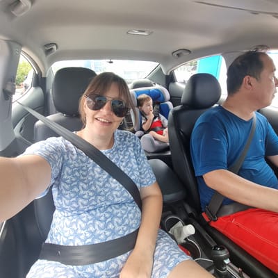 In Gangneung-si, South Korea, a family is captured in a selfie taken from the back seat of a car, likely during a road trip. The image shows a woman in the foreground wearing sunglasses and a blue and white patterned dress. She is smiling and appears to be the one taking the photo. Behind her, a toddler sits in a car seat, wearing a striped shirt and red shorts, looking towards the side. To the right of the woman, a man wearing a blue t-shirt and red shorts is visible, looking forward, presumably driving. The car interior is discernible, with black leather seats and seatbelts prominently featured, highlighting safety precautions. The background outside the car windows suggests an urban environment with buildings and trees, consistent with being in a city. The overall impression is a candid moment of a family enjoying a journey together.