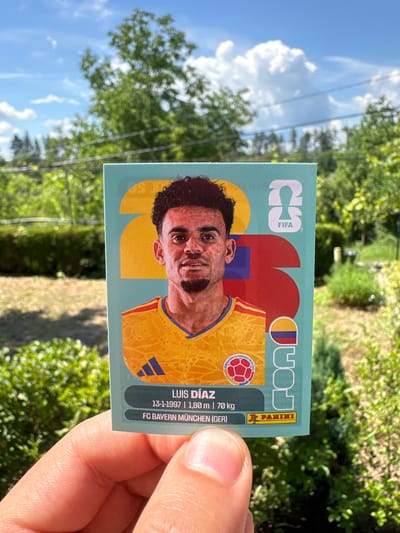 This is a close-up, outdoor, eye-level shot of a person's hand holding a trading card. The card features a portrait of a man with dark curly hair and a beard, wearing a yellow jersey with blue and red accents. He has a slight smile and his head is tilted. The card has text that includes "FIFA", "PANINI", and "LUIS DÍAZ". Below his name are his birthdate, height, and weight, followed by his club, "FC BAYERN MÜNCHEN (GER)". The background of the card is a light teal color with abstract shapes in red, yellow, and blue.
The background of the image is blurred but shows a sunny outdoor scene with green trees and a bright blue sky with white clouds. The lighting suggests it is daytime. The setting appears to be a natural environment, possibly a park or garden, given the lush greenery. The composition focuses on the trading card and the hand holding it, with the background out of focus.