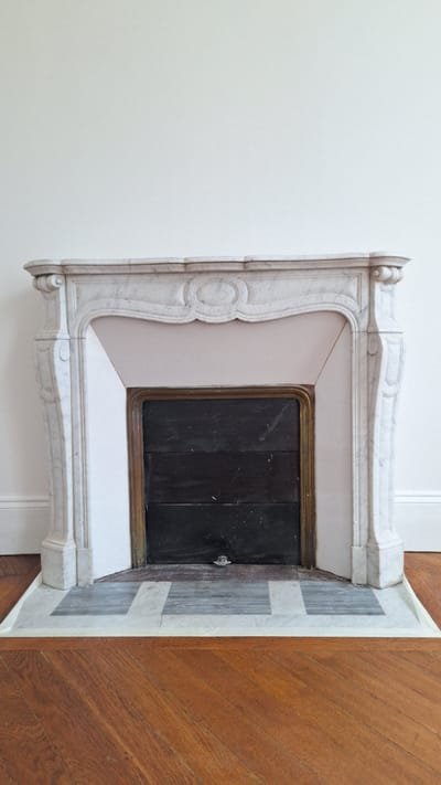 A marble fireplace is pictured in a room with a white wall and a wooden floor in Fontainebleau, France. The fireplace has an ornate, carved marble mantelpiece. Behind the mantelpiece, the firebox is covered by a dark, slatted panel, framed by a brass-colored metal. A small, metallic latch is visible in the center of the firebox panel. The hearth in front of the fireplace is made of marble tiles, with a white border and grey and white square tiles. The floor is made of polished wooden planks laid in a diagonal pattern. The scene is static, with no people or ongoing actions.