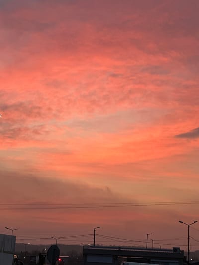 The image displays a wide-angle view of a sky at dawn or dusk over a terrestrial landscape in Plodovitovo, Bulgaria. The upper two-thirds of the frame are dominated by a sky exhibiting a color palette transitioning from deep orange and red at lower altitudes to pink and purplish-grey in the upper regions, indicative of scattered cloud formations. A small, bright, white crescent object is visible in the upper-left quadrant. The lower third of the image shows a silhouetted foreground composed of urban and semi-urban elements. Multiple streetlights with illuminated fixtures are positioned across the mid-ground, intersected by several parallel horizontal power lines. Partial structural details include a light-toned, block-built structure on the far left, and low-profile buildings with dark roofs, one featuring an external air conditioning unit, spanning the central and right portions of the foreground. A large, dark, circular object is centrally located in the lower frame. A dark band of distant vegetation delineates the horizon line. No human subjects or discernible interactions are present.