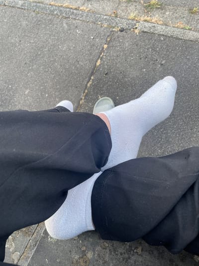 This image, taken in Wiehl, Germany, shows a close-up view of a person's legs and feet, seated outdoors on a paved surface. The person is wearing black pants and white socks. One foot is partially visible, wearing a light gray slip-on shoe. The ground appears to be a mixture of asphalt and concrete, with some sparse vegetation growing along the edges. The lighting suggests it is daytime, possibly late afternoon or early morning due to the angle of the shadows. There are no other people or objects clearly discernible in the frame.