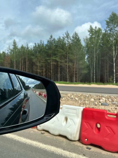 The image captures a view from a car's side mirror, reflecting a road construction site. In the foreground, red and white plastic barriers line the side of the road, suggesting ongoing work. Beyond the barriers, a dense forest of pine trees stretches across the horizon under a cloudy sky. The reflection in the mirror shows the car's rear, a paved road with white lane markings, and more red and white barriers further ahead, indicating the extent of the construction. The overall scene suggests a rural or semi-rural environment in the Задорʼеўскі сельскі Савет region of Belarus, characterized by a road undergoing maintenance or improvement amidst a natural wooded landscape. There are no people visible in the image.
