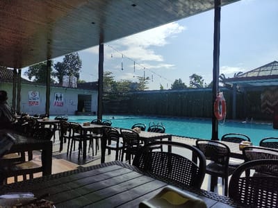The image shows a view of a swimming pool from a covered patio area. The pool is surrounded by a bamboo fence. There are several people swimming in the pool. The patio is furnished with black wicker chairs and tables. A life buoy is visible hanging on the support beam of the patio. A person is sitting at a table in the foreground, looking toward the pool. The sky is blue and clear with a few white clouds. The patio is covered with a wooden roof with dark wooden beams. Several warning signs are visible in the background, including one that reads "WARNING!". The location is a swimming pool in Gambiran, Indonesia, with coordinates -8.434 latitude and 114.18 longitude.