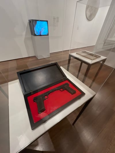 This image depicts an interior view of an art gallery or museum exhibit located in Cambridge, United States. The primary subject is a dark, open presentation case resting on a white rectangular display pedestal, enclosed within a clear acrylic vitrine. Inside the case, two dark brown, stylized pistol-shaped objects are set against a bright red, molded interior lining. The two pistols appear to be fused at their barrels, forming a single, horizontally oriented piece.
In the background to the left, an angled white pedestal supports a vintage-style cathode-ray tube (CRT) television displaying a dynamic, abstract image predominantly featuring blue and purple hues. Further back on the right, another white pedestal holds a light-colored, flat rectangular object contained within a second clear display case. A large, circular or oval, textured white artwork is partially visible on the far right wall, behind a clear partition. The gallery features plain white walls and a polished dark wood floor. No individuals are present in the frame.