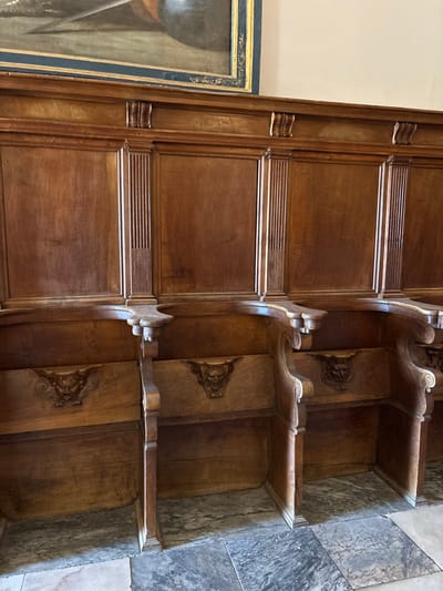 This image captures a section of ornate wooden choir stalls and paneling located within the Royal Monastery of San Lorenzo de El Escorial, Spain. The scene depicts an interior space, devoid of human subjects or direct actions.

The primary subjects are a series of interconnected, dark, polished wooden structures functioning as both seating and architectural paneling. The upper portion, acting as wainscoting, features rectangular wooden panels separated by vertical, fluted pilasters. A decorative wooden frieze with carved scroll-like elements runs along the top of this paneling. Above the frieze, a plain, light-colored wall is visible. In the upper left quadrant, a portion of a framed painting is present, displaying dark tones with what appears to be metallic weaponry (e.g., a sword, helmet) against a dark background. The painting's frame is dark blue with a golden inner border.

The lower portion consists of individual, high-backed wooden stalls. Each stall is defined by substantial, curved armrests and solid wooden dividers. Integrated into the upper part of each seat back, directly beneath the armrest, is a detailed, grotesque carved wooden face or mask. The floor is composed of large, polished stone tiles in varying shades of grey, white, and light brown, arranged in an irregular pattern. The environment is illuminated by ambient light, highlighting the intricate carvings and natural grain of the wood.
