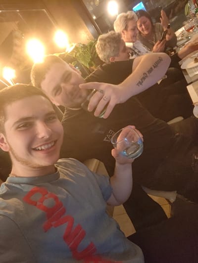 This image depicts an indoor social gathering, likely within a restaurant or bar setting in Kortrijk, Belgium. Five individuals are present. In the immediate foreground, a young male with short dark hair and light facial stubble wears a light blue t-shirt with red, partially legible text. He smiles directly at the camera, holding a clear, empty glass in his right hand. Behind him and slightly to the right, a second young male with short light-brown hair wears a dark t-shirt and smiles towards the foreground. He holds a glass containing a dark liquid in his left hand. A tattoo with inverted text is visible on his left forearm, and a small smiley face tattoo is on his right index finger. In the background, three women are seated at a table. One woman with dark hair is actively engaged with a smartphone held in her hands, her right hand raised as if gesturing or reacting, and she appears to be laughing. Two other women with short, light-colored hair are also seated at the table, facing generally towards the left. The table is set with plates and wine glasses, some containing red liquid. The environment features warm, artificial lighting that creates bright flares, and a potted plant with green foliage is visible. The seating appears to be dark-colored.