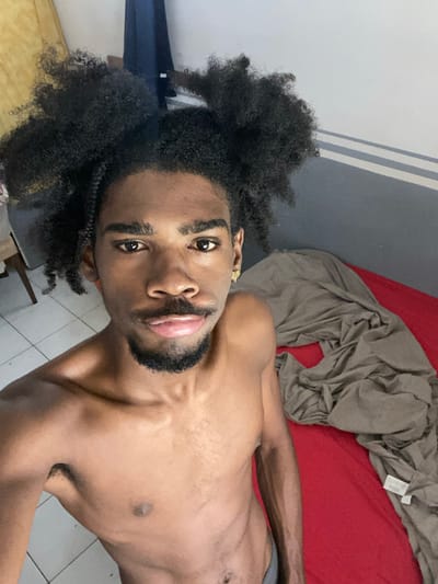 A shirtless, dark-skinned man with afro hair is lying on a red bed. A grey sheet is partially covering him. The background consists of a grey and white wall, and tiled flooring. There is no indication that the scene is specifically located in the city of Capesterre-Belle-Eau, France.