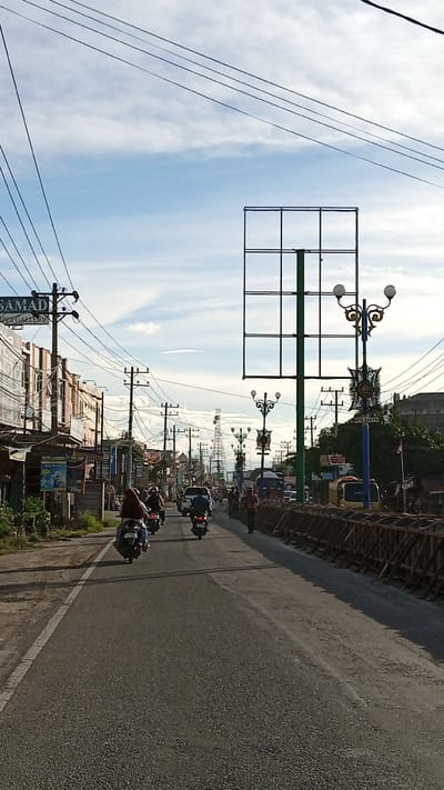 The image depicts a street scene in Geulanggang Teungoh, Indonesia, under a partly cloudy sky. The road is paved and has a white line marking the edge of the lane on the left side. Several motorcycles are visible on the road, with riders on them. Some are moving forward, while others appear to be stopped or moving slowly. There are buildings lining the left side of the street, some with signs, including one that reads "SAMADI PET GALLERY." Utility poles with numerous wires are prominent throughout the scene. On the right side of the road, there is a construction barrier made of wooden planks and supports. In the background, there are more buildings, trees, and telecommunication towers. The overall impression is that of a busy street in a semi-urban environment.