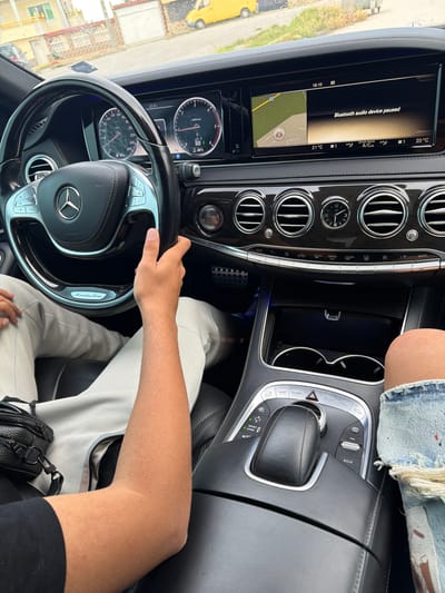 The media file displays the interior of a Mercedes-Benz car from a first-person perspective, likely the driver's. A person's right hand rests on the steering wheel, and their leg, wearing light-colored pants, is visible. A second person's leg, clad in distressed denim shorts, is partially seen on the right side of the frame. The car's luxurious black interior features polished wood trim, chrome accents, and leather seating.
The car appears to be stationary, with the infotainment screen showing "Bluetooth audio device paused" and a navigation map. The time displayed is 18:10. The instrument cluster indicates the car is in "P" (Park) and shows a tachometer, speedometer (up to 260 km/h), and other readings like "90℃" and "Trip 25%." The climate control displays individual temperature settings of 21°C and 20°C.
Through the front window, a street scene is visible with buildings and a yellow vehicle. Rain droplets on the window suggest wet weather or recent rain. The navigation map shows local street names such as "Ул. Цар Симеон" and "Ул. Драгомир," consistent with the location context of Dragor, Bulgaria.
Visible text includes: "Mercedes-Benz," "AIRBAG," "18:10," "Bluetooth audio device paused," "21°C," "20°C," "AUTO," "MENU," "NAVI," "RADIO," "MEDIA," "TEL," "ON," "VOL," "ECO," "SPORT," "COMFORT," "P," "D2 E 6," "90℃," "Trip," "25%," "x1000/min," "Ул. Цар Симеон," and "Ул. Драгомир."