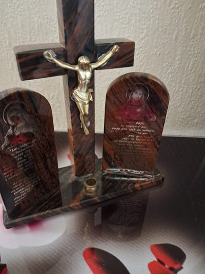 This image, taken in Gavar, Armenia, showcases a religious devotional object crafted from polished, dark reddish-brown stone with lighter veining. The central element is a crucifix featuring a gold-colored figure of Jesus Christ mounted on a cross.
Flanking the cross are two upright, rounded rectangular stone plaques. The upper plaque displays an etched image of a religious figure, likely Jesus Christ or a saint, characterized by a halo and a red garment or heart symbol. Below this figure, Armenian text is inscribed. The text includes phrases like "Դու, տիրուհի," (You, Our Lady/mistress) and parts of an invocation: "աղաչում, անարյան բարեխոսիր ծ համար և Միածին Ո փորձու" (pray, intercede without blood for you and Only-Begotten O trial), suggesting a prayer, possibly to the Virgin Mary or a female saint.
The lower plaque, partially visible, also features an etched image and Armenian text. This text is identifiable as the beginning of the Lord's Prayer: "Հայր մեր, որ երկնքում ես. սուրբ թող լինի քո անունը. և թող լինի երկրի չպես որ երկնքում է. սար հանապազօրյա հացը տուր մեզ այսօր. h song մեզ մեր պարտքերը, - և մենք ենք թողնում" (Our Father, who art in heaven, hallowed be thy name. Thy kingdom come, thy will be done, on earth as it is in heaven. Give us this day our daily bread; and forgive us our trespasses, as we forgive those who trespass against us).
A small brass-colored cylindrical fitting is visible near the base of the cross. The background to the left is a light, textured wall, while the right side reveals a darker surface with blurred red abstract shapes, possibly part of a floral print. The overall composition conveys a solemn and reverent atmosphere, typical of religious artifacts.