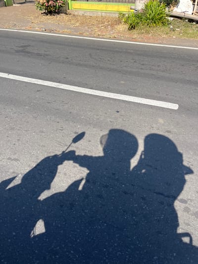 The image captures a shadow of two individuals on a paved road in Purworejo, Indonesia, with a white line marking the lane. The shadows suggest they are riding a motorcycle, with one person in front and another behind. The foreground is dominated by these silhouettes cast by the bright sun, which also illuminates the asphalt and the dry grass and foliage on the side of the road. A yellow-painted wall and a green fence are visible in the background, indicating an urban or semi-urban setting. The overall scene is a snapshot of everyday life on a sunny day in this Indonesian city.