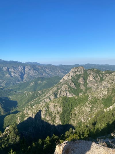 The image depicts a panoramic view of a rugged, mountainous landscape under a clear blue sky. The terrain is characterized by steep, rocky slopes covered in dense green vegetation, primarily trees and shrubs. In the foreground, a rocky outcrop with sparse vegetation is visible, casting a long shadow across the scene. The middle ground features a prominent, jagged mountain peak with interspersed patches of green foliage clinging to its rocky surfaces. Beyond this, a series of receding mountain ranges stretch towards the horizon, becoming progressively softer and more muted in color. A deep valley or ravine is visible to the left, with a winding path or riverbed cutting through the lush green slopes. The lighting suggests it is either early morning or late afternoon, as evidenced by the long shadows. There are no people or man-made structures immediately apparent in the visible portion of the image. The overall impression is one of a remote and wild natural environment.