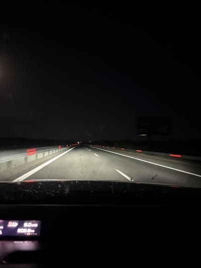 The image shows a view from inside a vehicle driving on a highway at night. The road is illuminated by headlights, with white lines marking the lanes and red reflectors along the side. The sky is dark, indicating nighttime. In the foreground, a digital display from the vehicle shows a speed of 6.0 and a distance of 202.5. No people are visible in the image. The scene appears to be on a highway outside of Malko Tranovo, Bulgaria, given the context provided.