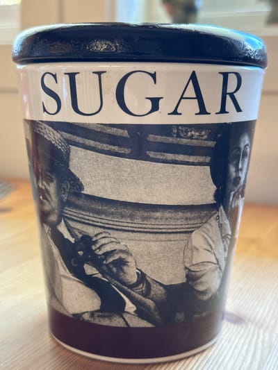 A close-up, eye-level shot shows a ceramic sugar canister with a black lid. The canister is white with black lettering that reads "SUGAR" in uppercase serif font across the top. Below the text, a black and white vintage photograph wraps around the canister. The photograph depicts two people in what appears to be a car. The person on the left wears a hat and a collared shirt, with a cigarette in their mouth. Their hands are visible, holding onto a steering wheel. The person on the right, partially obscured, has a surprised expression with their mouth open. The background of the photograph features striped upholstery. The canister sits on a light-colored wooden surface, possibly a table. The lighting is natural and casts a warm glow. The background is blurred, suggesting an indoor setting. There is no information to suggest this image is located in the city of Valby, Norway.