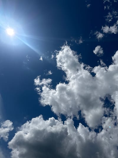 This media file depicts a clear blue sky with scattered white cumulus clouds, some of which are darker and appear to be gathering, suggesting a dynamic weather pattern. The sun is prominently visible in the lower left quadrant, emitting bright rays that create a lens flare effect across the image. The overall impression is one of a sunny day, possibly with some approaching weather.
The scene is an outdoor, natural setting, likely captured from a ground-level perspective looking upwards. There are no visible people or man-made objects, focusing the viewer's attention entirely on the sky and atmospheric conditions. The bright sun and the contrast between the deep blue sky and the white clouds indicate it is likely daytime, possibly midday or afternoon given the sun's position and intensity. The presence of various cloud formations could suggest a transitional period in the weather, perhaps leading to or from rain.
The image evokes a sense of openness and the vastness of the sky. The composition, with the sun in the corner and clouds filling the rest of the frame, draws the eye across the expanse. No text is visible in the image. The given location context of Мазалаўскі сельскі Савет, Belarus, places the scene in a rural area of Belarus, further reinforcing the natural, open-air environment.