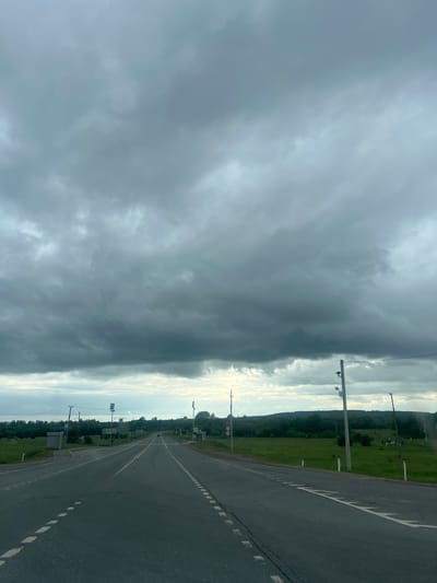 The image captures a view from a vehicle traveling on a paved road in Гремяча, Russia. The road is asphalt with white dashed lines indicating lanes and is bordered by grassy shoulders. The surrounding landscape is rural, with green fields and a dense line of trees in the distance, forming a horizon. The sky is heavily overcast with dark, heavy clouds, suggesting impending rain or stormy weather. Several utility poles with electrical wires and street lights are visible along the roadside. Road signs are present, including what appear to be pedestrian crossing signs and speed limit signs. A small bus stop shelter is also visible on the left side of the road. No people or animals are clearly discernible in the scene, and the overall atmosphere is somber due to the weather conditions.