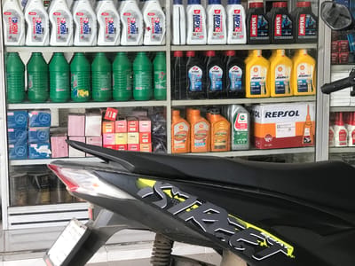 This is an indoor shot of a motorcycle parked inside or in front of a shop in Rambot, Indonesia. The shop shelves behind it are stocked with various brands of motorcycle oil and lubricant products.  Specific brands visible include Repsol, Shell Advance, Castrol Activ, Motul Scooter LE, UDSOL, Honda MPX2, NBX2, and GGI. The motorcycle is a black "Beat" model.  Boxes of parts and oil cans are also visible on the shelves.