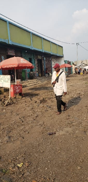 The image depicts a street scene in Buzove, Democratic Republic of the Congo. A male subject, wearing a red baseball cap, sunglasses, a light-colored long-sleeved shirt, dark trousers, and sandals, walks from right to left across the unpaved, uneven dirt road in the mid-ground. A black strap from a bag or equipment crosses his chest. To the left of the frame, a red umbrella prominently displaying the "Airtel" logo shades a small, red, mobile vendor cart and a white sign board on a stand with illegible script. Behind these objects, a row of multi-story buildings with weathered facades, featuring ground-level shops with various doors and shutters, lines the street. The upper section of these buildings exhibits a light blue base supporting a light green horizontal band. A partial "STARIS" sign is visible above one shop. Further down the street on the left, another red Airtel umbrella is discernible. The ground consists of dry, compacted brown dirt with visible ruts and scattered debris, including a dark cylindrical object in the foreground. In the far background on the right, an area of informal structures constructed from various materials is visible, along with faint human figures. A utility pole with overhead lines is present on the right side of the scene. The sky is overcast and uniformly light grey.