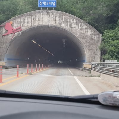 The image shows the entrance to the Jangpyeong 2 Tunnel in Yongpyeong-myeon, South Korea. A red silhouette of a skier is visible on the left side of the tunnel entrance. Above the tunnel entrance, a blue sign reads "장평2터널" which translates to "Jangpyeong 2 Tunnel". The tunnel itself is dark, with lights lining the ceiling. Orange traffic cones and yellow lines mark the roadway leading into the tunnel. The surrounding environment appears to be mountainous with lush green trees. The photo seems to be taken from inside a vehicle, possibly a car, as the dashboard is partially visible in the foreground.