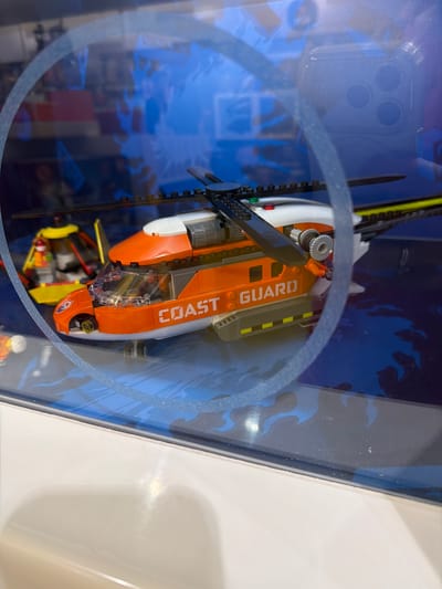 A LEGO Coast Guard helicopter is displayed inside a clear plastic box. The helicopter is predominantly orange and white, with "COAST GUARD" printed in white letters on the side. It has a black rotor and tail fin, and a clear cockpit. The background of the display box features a blue, wave-like graphic, suggesting a marine or water-related theme. The helicopter appears to be a model, possibly part of a toy set. The lighting suggests an indoor setting, likely a store or display case. There is a faint reflection on the plastic, showing what appears to be a phone case with three circular camera lenses. The image is oriented sideways.