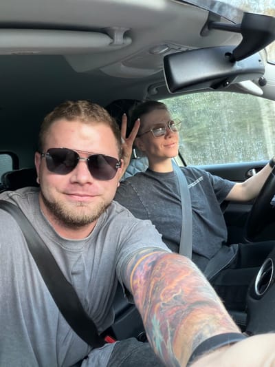 Two people are in a vehicle. A person wearing sunglasses is in the foreground. Another person, wearing glasses and holding up a peace sign, is in the background. Both are wearing seatbelts. The setting is inside the vehicle, seemingly on a rainy day. There is no direct visual evidence to confirm the location is Bol'shaya Sosnova, Russia.