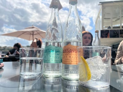 This image depicts an outdoor tabletop scene in Porto Cristo, Spain, under a partly cloudy sky. In the immediate foreground, two clear glass bottles of "Font Major" mineral water stand on a dark, reflective surface, likely a table. The left bottle displays a teal label identifying it as "Natural Mineral Water" and appears full. The right bottle has an orange label for "Sparkling Mineral Water" and is also full. To the left of the bottles, a clear glass contains water, approximately half-full. To the right, another clear glass holds several ice cubes and a large yellow lemon wedge, with discernible effervescence in the clear liquid.
In the blurred midground, several individuals are seated. On the left, a person with dark hair is partially visible under an open, light-colored patio umbrella, with additional indistinct figures behind them. On the right, a woman with dark hair is visible behind the bottles, looking towards the camera, with her right arm and shoulder also in view. The background reveals a blurred body of water, consistent with a harbor, and indistinct architectural elements, including a glass-paneled structure on the far right. The scene suggests an outdoor cafe or restaurant setting.