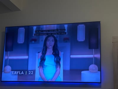 A large, wall-mounted flat-screen television dominates the center of the frame, displaying a scene from a television program. The screen features a young woman, identified by an on-screen graphic as "LAYLA | 22," centered in a studio-like setting. She has long dark hair and is wearing an off-the-shoulder light blue dress, with her mouth slightly open. Closed captions are visible at the top of the screen, stating, "I'm not really sure why Whitney would consider not going to this." The background on the television screen is minimalist, with blue ambient lighting illuminating vertical rectangular panels, dark vertical panels, a horizontal light bar, and spherical objects on pedestals.

The television is mounted on a light-colored wall. A blue LED strip light is affixed to the bottom edge of the TV, casting a blue glow downwards onto the wall. To the right of the television, a section of light wood-grain furniture, possibly a cabinet or wardrobe, is partially visible. The ceiling, painted a light color, is visible in the upper portion of the image. The immediate environment is dimly lit, with the television screen and its integrated blue LED strip providing the primary light source, imparting a blue cast to the surroundings.